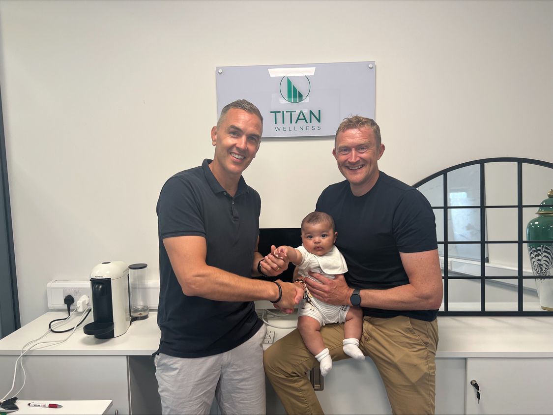 Titan Wellness co-founders Niall Ronan and Paul McManus shaking hands in front of the Titan Wellness sign, with a baby between them
