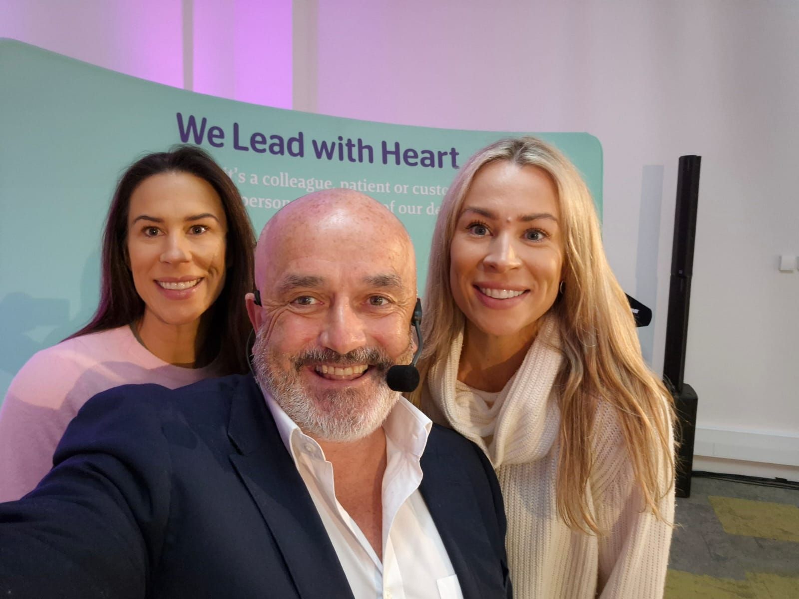 A speaker with a headset microphone smiling alongside two colleagues in front of a "We Lead with Heart" event banner