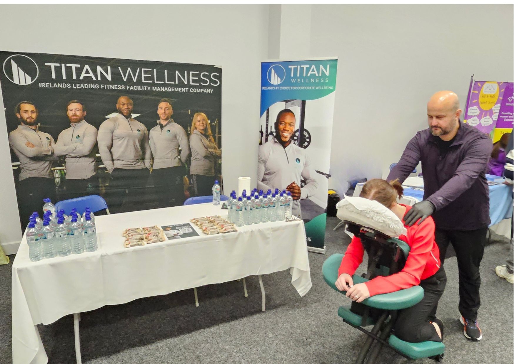 A man giving a woman a seated chair massage at a corporate wellbeing event, with Titan Wellness banners in the background