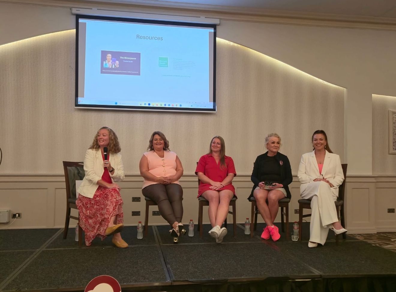 Panel of five women speaking at a female health and menopause awareness event, with a "Resources" presentation slide on screen behind them
