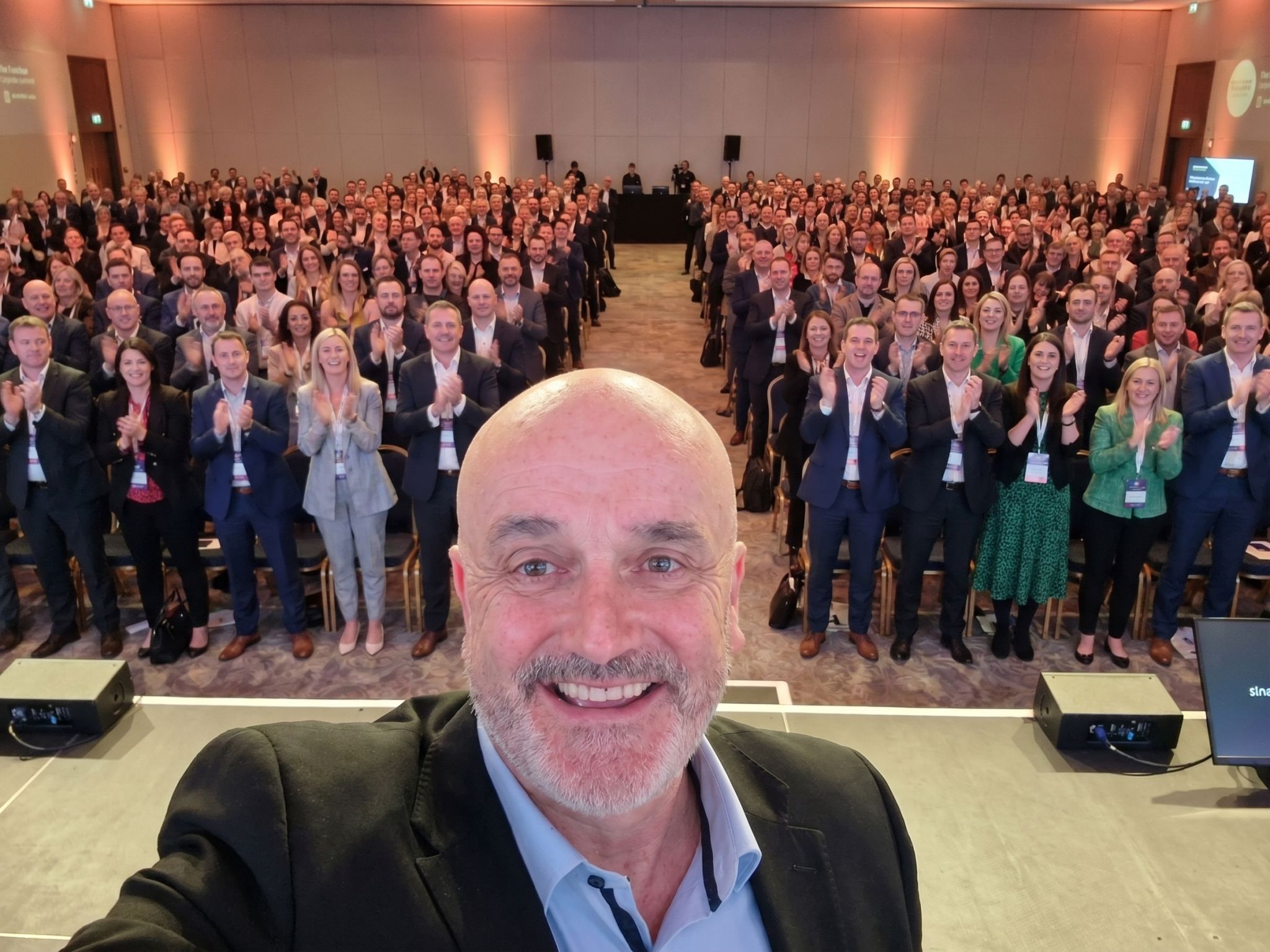 Speaker taking a selfie on stage in front of a large conference audience applauding