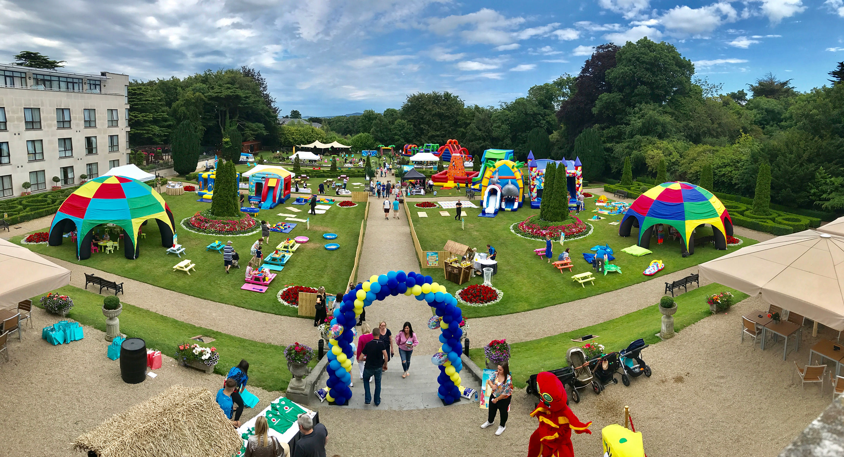 Aerial view of a corporate games event in hotel gardens with colourful inflatable domes, bouncy castles, picnic tables and a balloon arch
