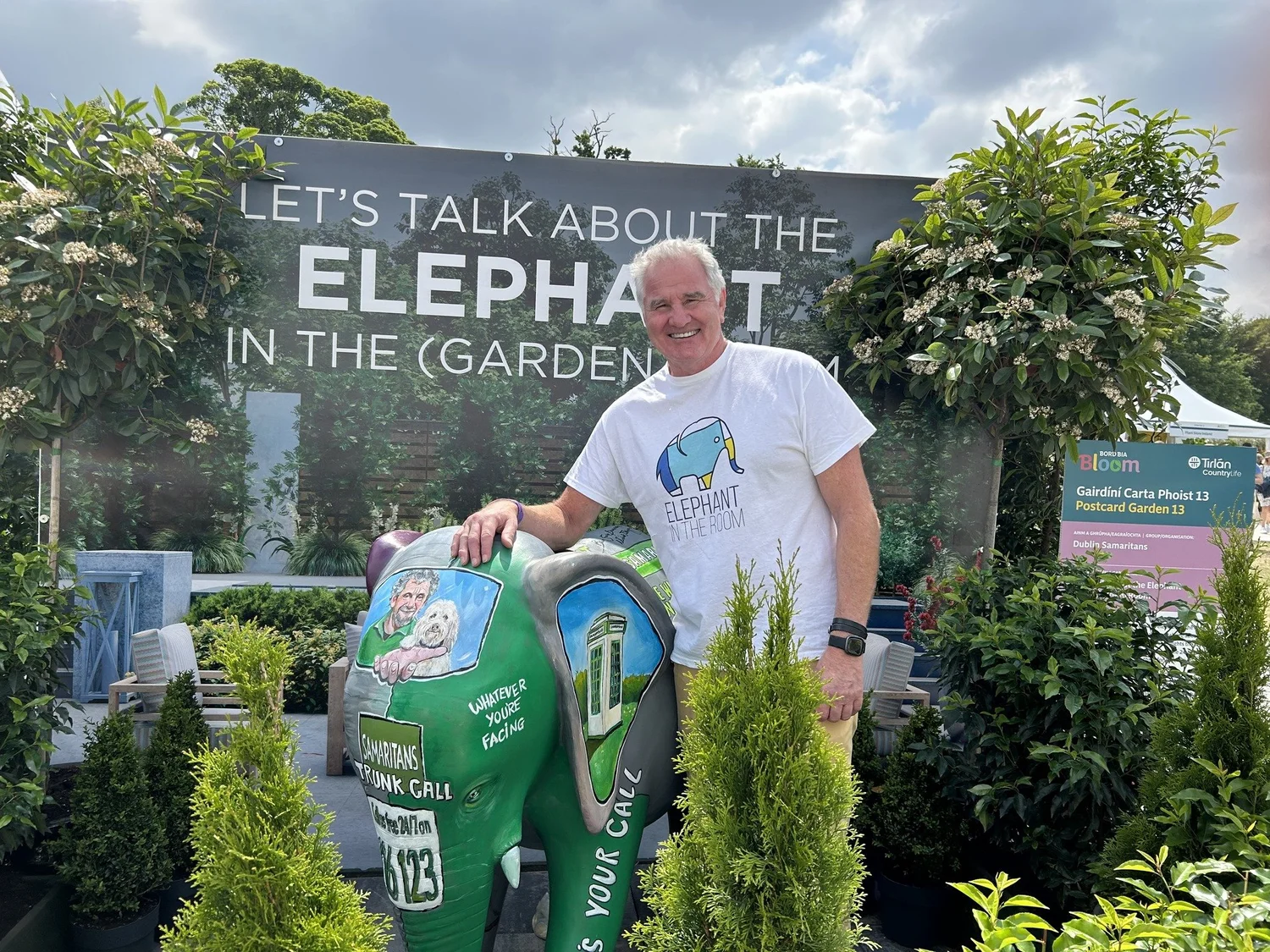 A keynote speaker smiling beside a hand-painted elephant statue at a Bloom garden festival wellbeing display