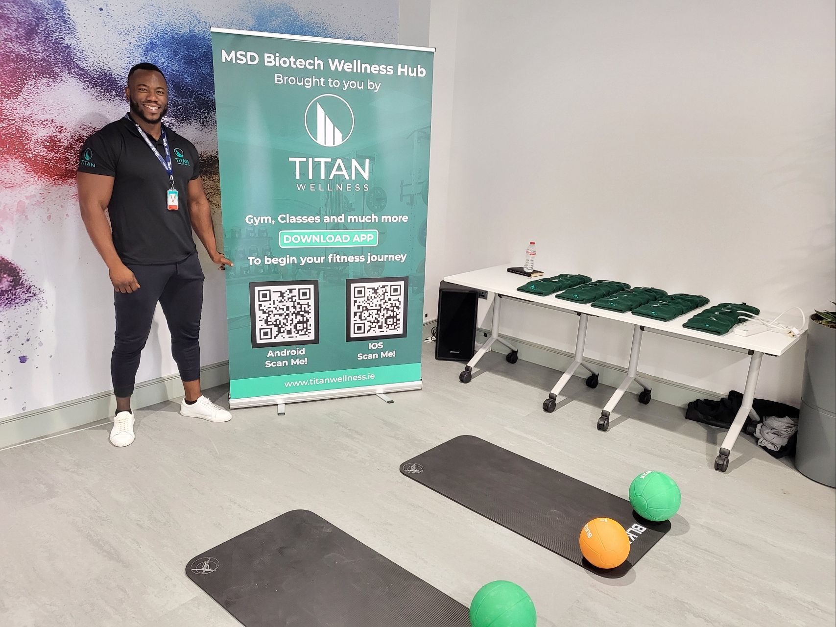 An Titan Wellness Fitness Facility Manager beside a Wellness Hub banner, with gym equipment, mats and medicine balls set up for a session.